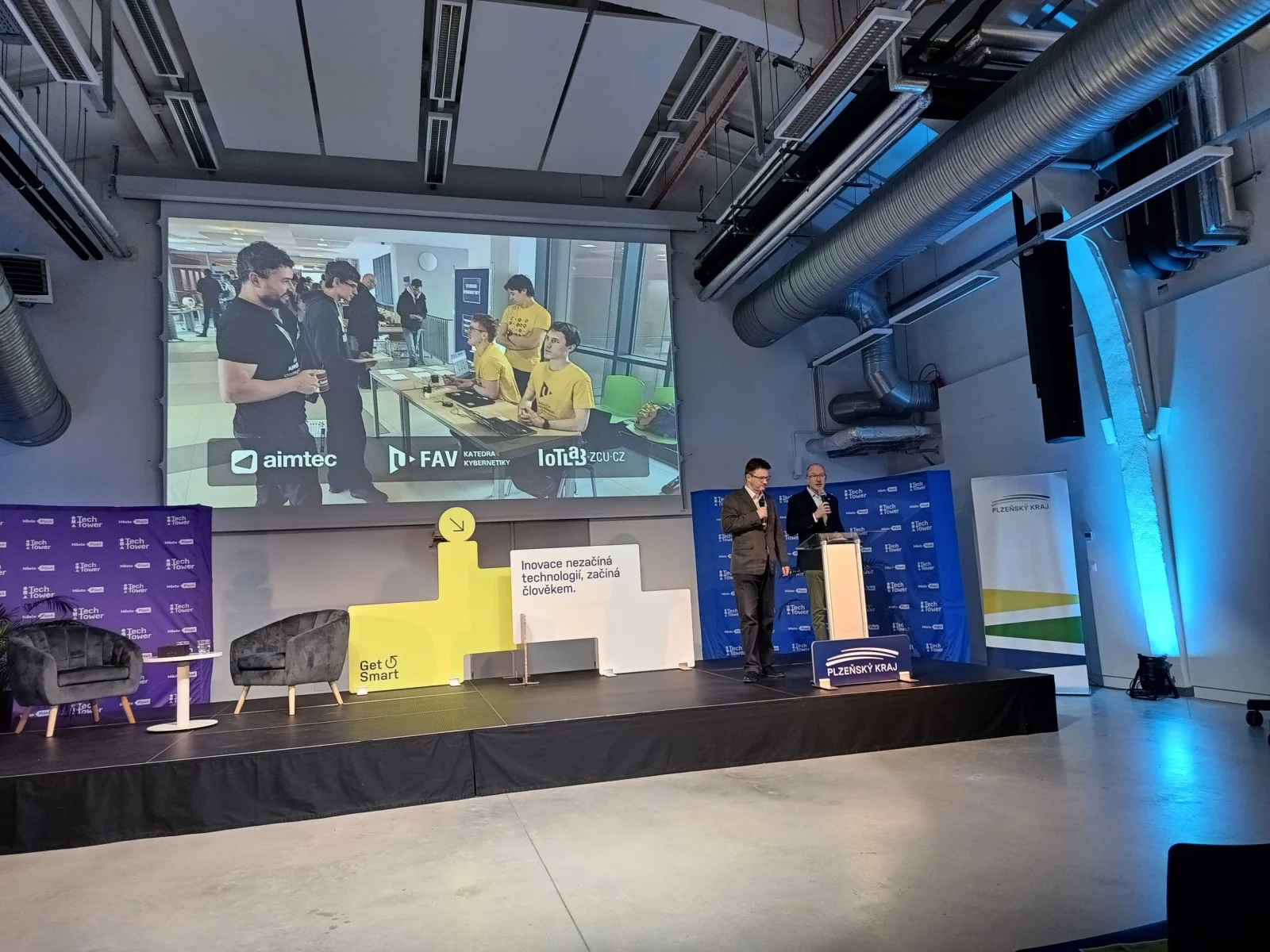 Two speakers holding microphones on stage at TechTower, with a student project photograph and Aimtec, FAV and IoT Lab logos projected behind them